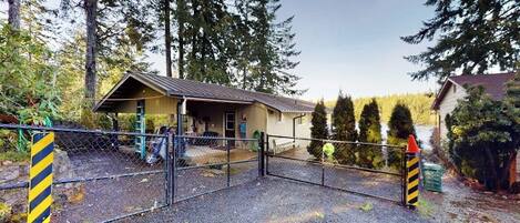 A fenced property with a small building, gravel ground, and tall trees in the background.