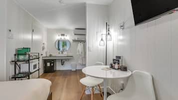 A modern hotel room with a round dining table, white chairs, a small kitchenette, a bathroom with a mirror, and a flat-screen TV mounted on the wall.