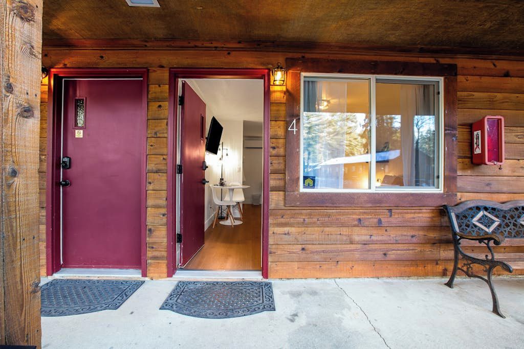 L’entrée d’un chalet en bois, avec une porte rouge, un banc et une fenêtre donnant sur une forêt.