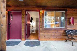 L’entrée d’un chalet en bois, avec une porte rouge, un banc et une fenêtre donnant sur une forêt.