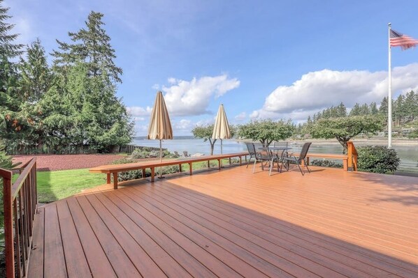 Una terraza de madera con mobiliario exterior, una bandera y vistas a una masa de agua.