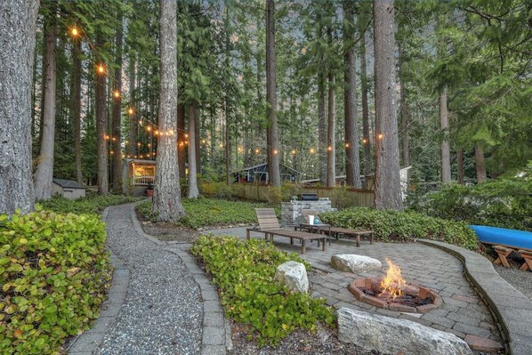 A forested outdoor area with a fire pit, seating, and string lights. - Skiing 1BR Lakefront | Dock | Deck | Firepit (Fall City)