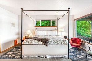 Un lit à baldaquin avec une couvre-lit à motifs floraux, un fauteuil rouge et une vue sur la verdure par la fenêtre.