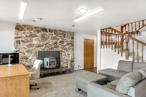 Un salon avec un mur en pierre, une cheminée, un canapé gris, un bureau en bois et un escalier.