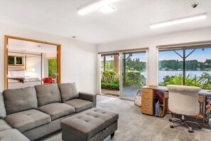 A modern living room with a grey sofa, a small ottoman, a desk with a chair, and large windows offering a view of a lake and greenery. - 4BR Lakefront - Dock - Fireplace - Balcony (Olympia)