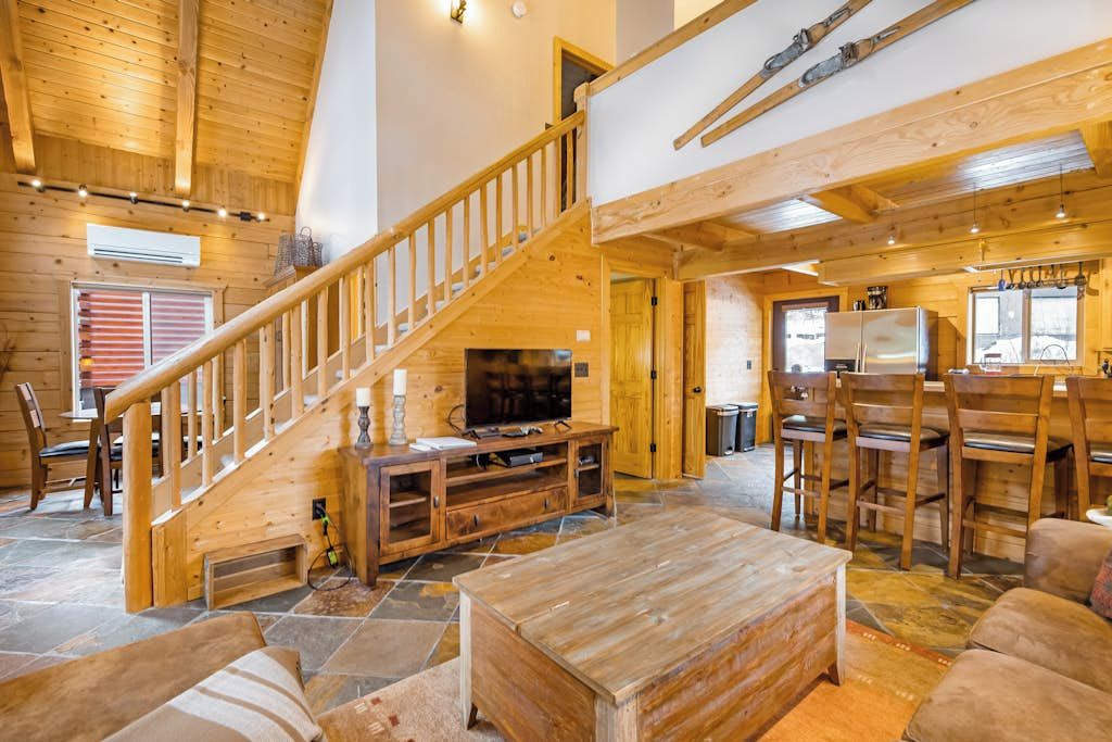 A wooden interior with a staircase, a television, a dining area, and a stone floor.