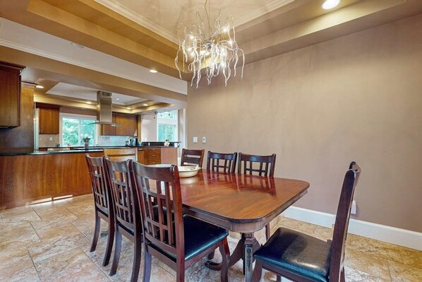A dining area with a wooden table and chairs, a chandelier, and a kitchen in the background.