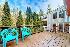 A wooden deck with two turquoise chairs, a metal railing, and a view of tall evergreen trees.