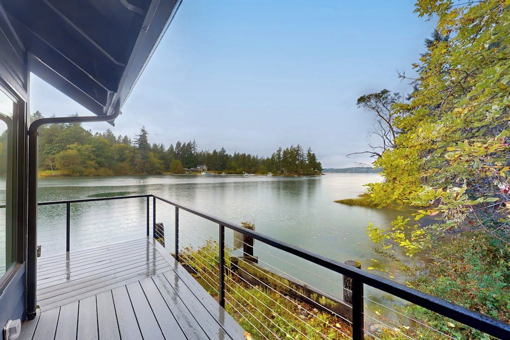 Een houten terras met een metalen hekwerk met uitzicht op een rustig meer, omringd door bomen en een heldere lucht.