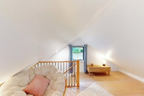 Un dormitorio acogedor en el ático, con techo inclinado, un sillón cómodo, una mesita de noche con lámpara y vistas a través de una ventana.