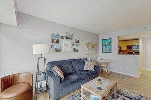 A modern living room with a sofa, coffee table, and lamp. There are framed pictures on the wall and a kitchen in the background. - 1 BR downtown condo with shared pool and hot tub (Seattle)