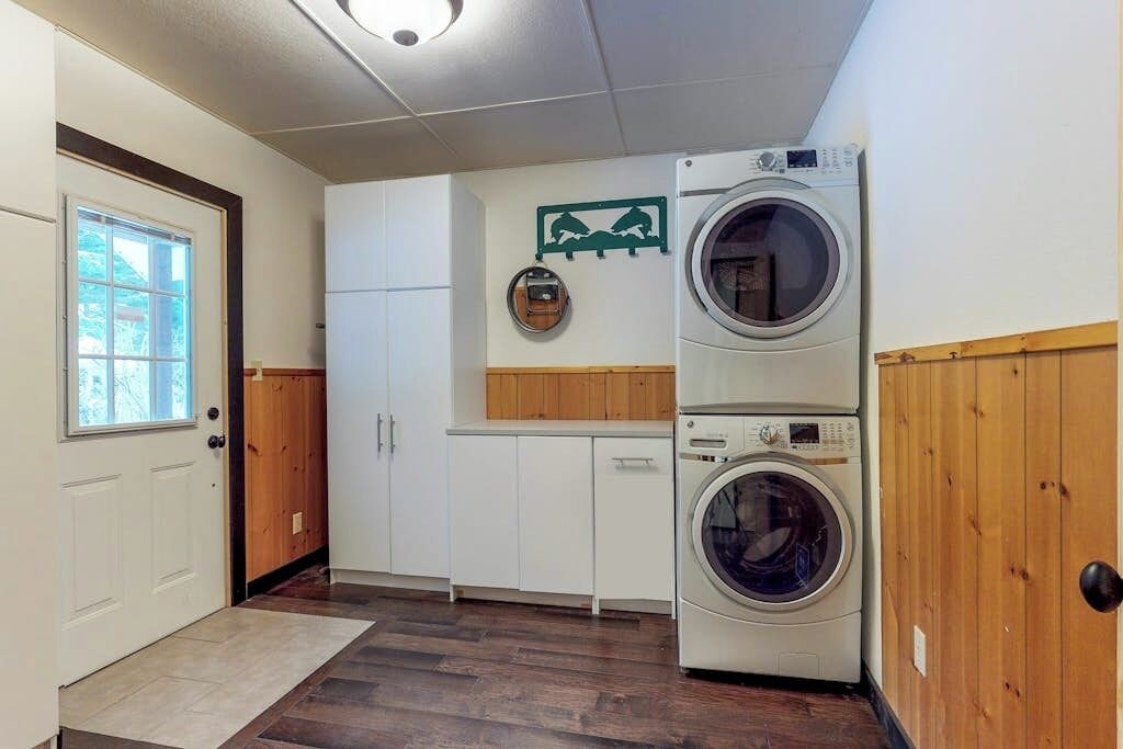 A laundry room with two stacked washing machines, wooden cabinets, a door with a window, and a wall-mounted drying rack.