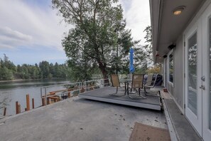A deck with outdoor furniture overlooking a lake, a covered area with a blue umbrella, and a building with large windows.