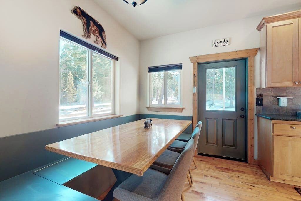 A kitchen with a wooden table, built-in benches, a door with a 'Family' sign, and windows with a view of trees.