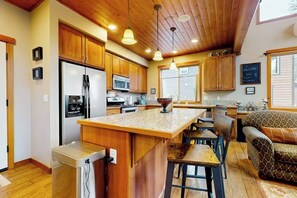 A kitchen with wooden cabinets, a central island, bar stools, and a dining table. - 3BR all-season cabin with deck - walk to the lifts (Snoqualmie Pass)