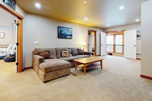 A living room with a sofa, ottoman, and coffee table. There is a painting on the wall and a door leading to another room. - 3BR all-season cabin with deck - walk to the lifts (Snoqualmie Pass)