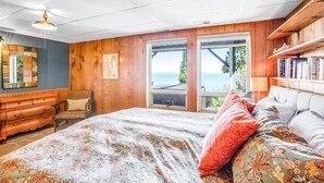A bedroom with a large bed, wooden walls, a dresser, a chair, and a view of the outdoors. - Impeccable 3BR Oceanview Dog Friendly | Hot Tub (Port Ludlow)