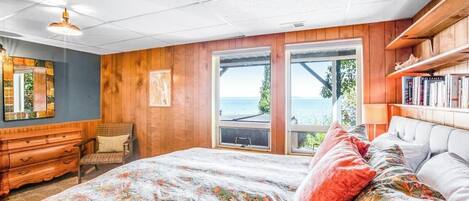 A bedroom with a large bed, wooden walls, a dresser, a chair, and a view of the outdoors.