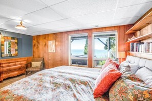 A bedroom with a large bed, wooden walls, a dresser, a chair, and a view of the outdoors.