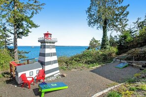 A lighthouse with a red roof, a small adjacent building, red chairs, and a green board game on a gravel area with a view of the sea.