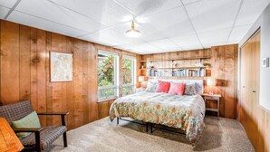 A bedroom with a wooden paneled wall, a bed with a floral bedspread, a bookshelf, and a window with a view of greenery.