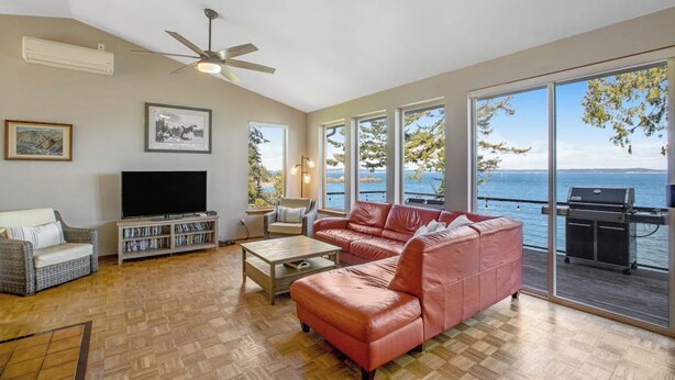 A living room with a television, sofa, and coffee table, large windows with a view of the sea, and a ceiling fan. - Impeccable 3BR Oceanview Dog Friendly | Hot Tub (Port Ludlow)