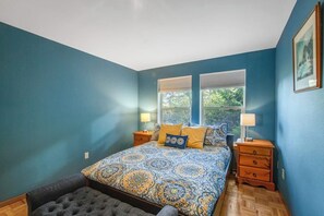 A bedroom with a patterned bedspread, two bedside tables with lamps, a bench, and a window with blinds. - Impeccable 3BR Oceanview Dog Friendly - Hot Tub (Port Ludlow)