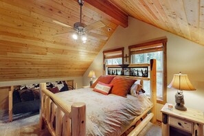 Ein Schlafzimmer mit schrägem Dach, einem Bett mit Holzrahmen, einem Nachttisch und einem Fenster mit Blick auf Bäume.