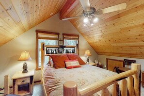 Quarto aconchegante com teto inclinado, cama com colcha florida, duas luminárias de cabeceira e janela com vista para árvores.