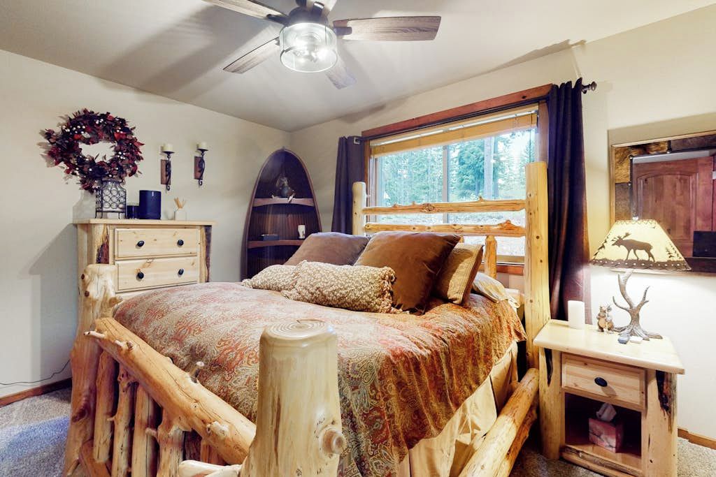 Une chambre à coucher avec un lit en bois, un ventilateur de plafond, une fenêtre avec des rideaux, une commode et une table de chevet avec une lampe.
