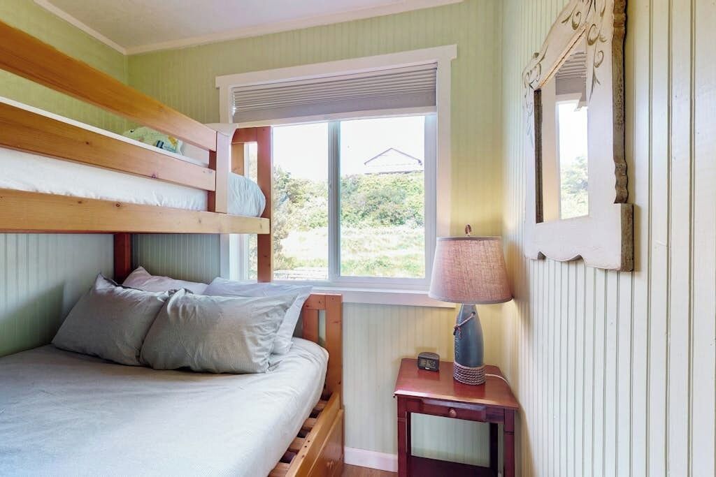 A room with a bunk bed, a bedside table with a lamp, and a window with a view of greenery.