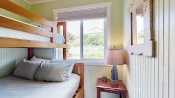 A room with a bunk bed, a bedside table with a lamp, and a window with a view of greenery.