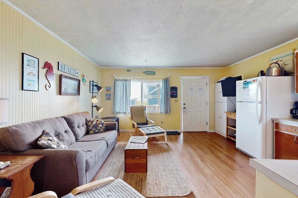 A compact living space with a sofa, a small table, a chair, a kitchen area with a refrigerator, and a window with curtains.