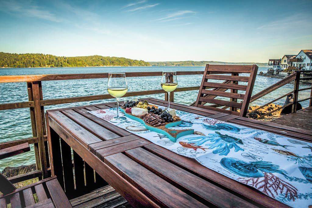 Une terrasse en bois avec une table dressée pour un repas, donnant sur une étendue d’eau et une zone boisée.