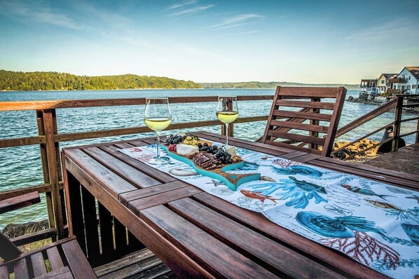 Eine Holzterrasse mit gedecktem Tisch, mit Blick auf ein Gewässer und einen bewaldeten Bereich.