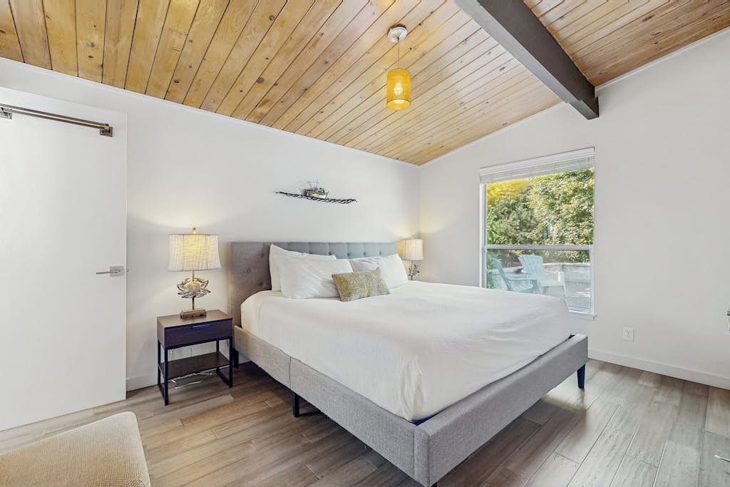 Een slaapkamer met een groot bed, houten balken in het plafond, een nachtkastje en een raam met uitzicht op groen.
