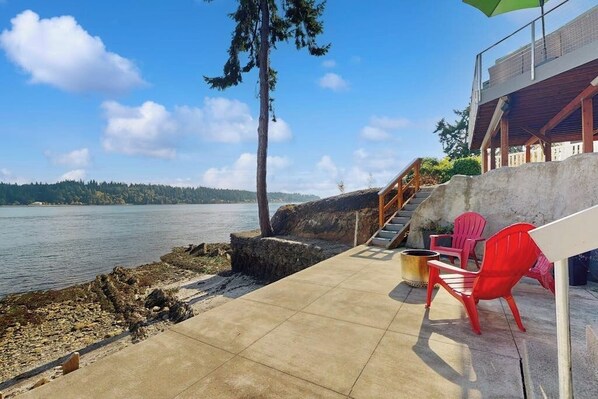 A concrete deck with a view of a lake, red chairs, and a staircase leading to a building. - Modern Waterfront Home with Amazing Views (Port Orchard)