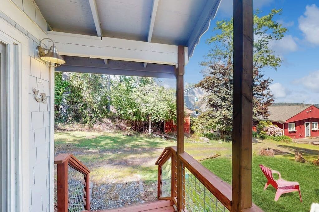 A covered porch with a wooden railing, a red chair, and a view of a grassy yard with trees and a red building in the background.