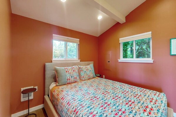 A bedroom with a bed, two windows, a bedside table, and a wall-mounted shelf.