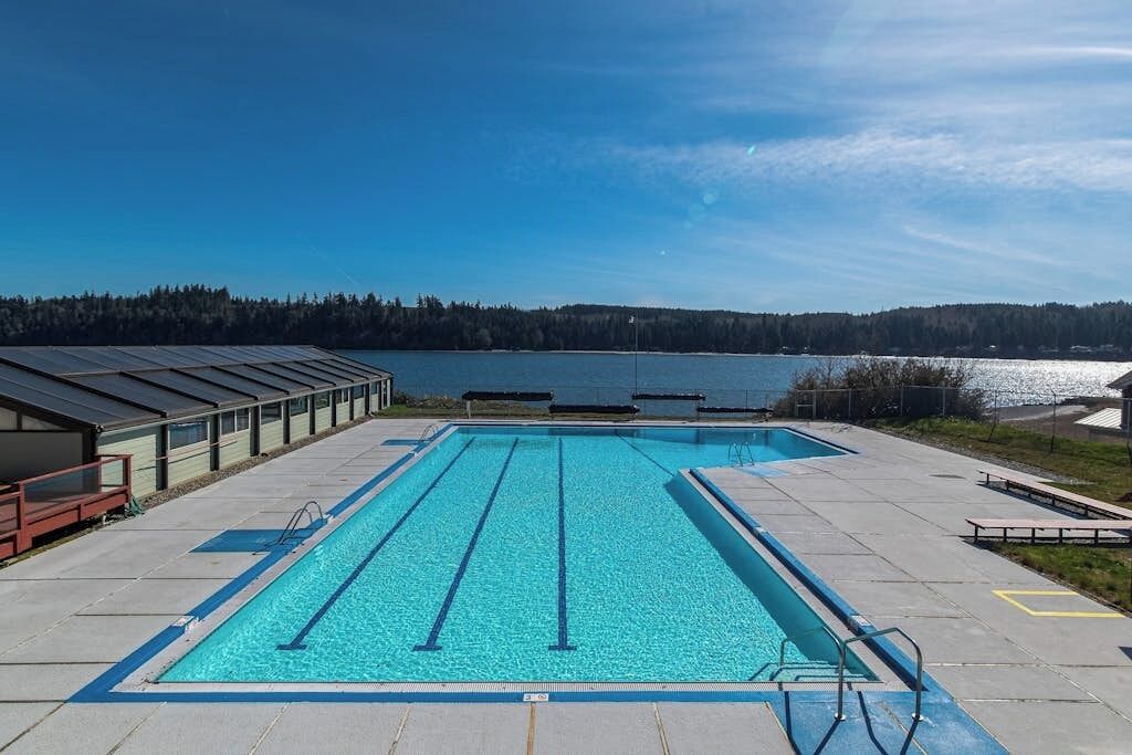 A rectangular swimming pool with lane dividers, surrounded by a concrete deck, with a view of a lake and forested hills in the background.