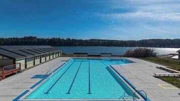 A rectangular swimming pool with lane dividers, surrounded by a concrete deck, with a view of a lake and forested hills in the background.