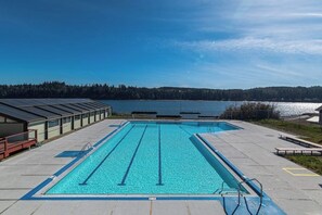 A rectangular swimming pool with lane dividers, surrounded by a concrete deck, with a view of a lake and forested hills in the background.