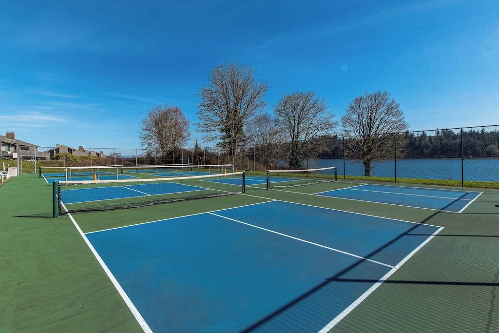 An outdoor tennis court with multiple courts, surrounded by trees and a body of water.