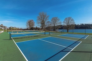 An outdoor tennis court with multiple courts, surrounded by trees and a body of water.