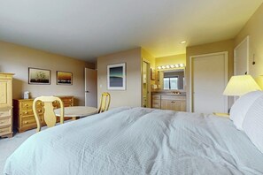 A hotel room with a bed, a desk, a chair, a vanity with a mirror, and a lamp.