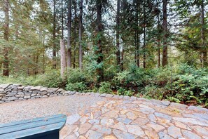 A stone patio with a wooden bench, surrounded by a stone wall and dense forest.