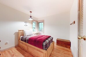 Ein Schlafzimmer mit einem hölzernen Bett, einer roten Decke, einem Nachttisch, einer Nachttruhe, einer Wanduhr und einem Fenster.