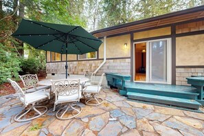 Eine Außenpatio mit Steinböden, einem runden Tisch, weißen Korbstühlen, einem grünen Sonnenschirm und einer Holzterrasse, die zu einem Gebäude mit einer Steinfassade und großen Fenstern führt.