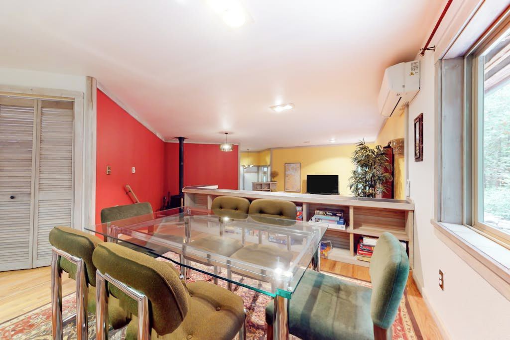 A dining area with a glass table and chairs, a red accent wall, and a window with an air conditioner.