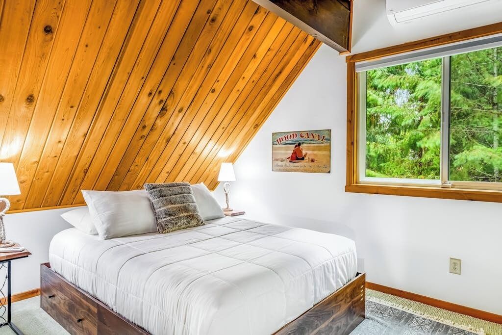 Ein Schlafzimmer mit geneigter Decke, einem großen Fenster mit Blick auf Bäume, einem Bett mit weißer Bettdecke und einem Poster an der Wand.